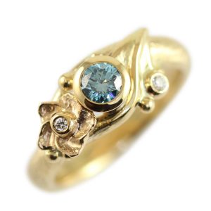 Smuk forlovelsesring i guld med dragehale og blomst samt en stor Ocean Blue diamant og små hvide diamanter